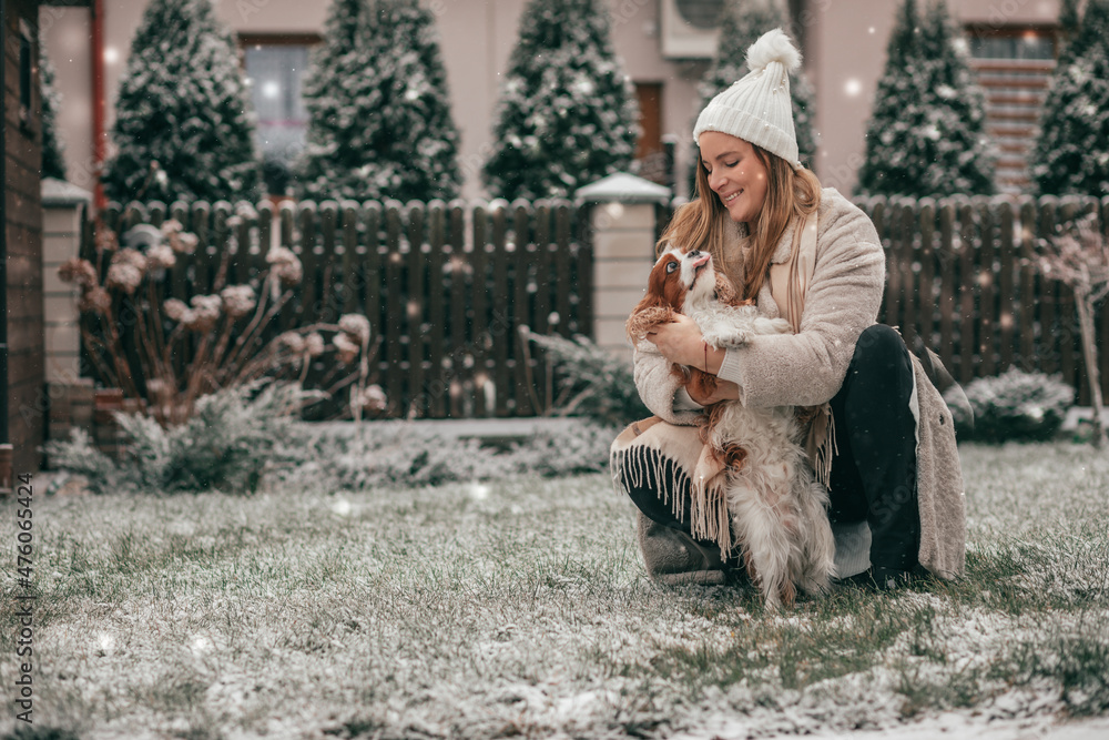 Charming young woman in white knitted hat is playing in snowy garden with dog. Cavalier King Charles spaniel. Winter landscape. New Year's card. Pets. Wonderful moments . Lot of happiness. Charming young woman in white knitted hat is playing in snowy garden with dog. Cavalier King Charles spaniel. Winter landscape. New Year's card. Pets. Wonderful moments . Lot of happiness.