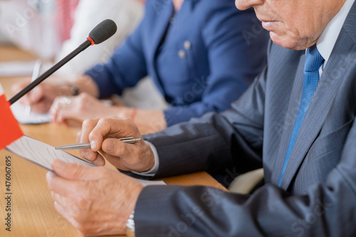 Hands of mature delegate pointing at document in clipboard while making report by table against row of foreign colleagues