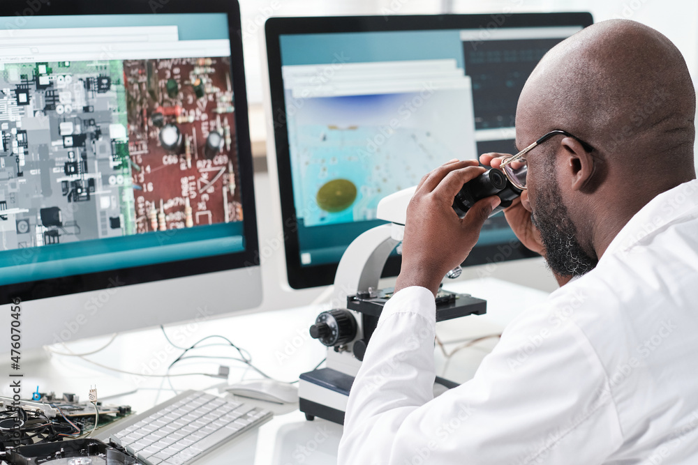 Young man of African ethnicity looking in microscope while sitting in front of computer screens with magnified microchip Young man of African ethnicity looking in microscope while sitting in front of computer screens with magnified microchip
