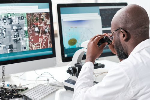Young man of African ethnicity looking in microscope while sitting in front of computer screens with magnified microchip