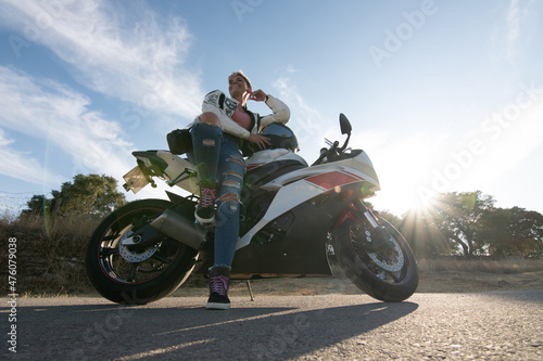 young pretty woman dressed in motorcycle equipment smiling leaning on her road motorcycle. sunset in the background