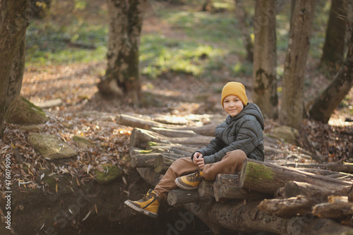 A cute blond boy in the spring forest is sitting on the bridge of the river. The boy looks into the frame. The concept of life after quarantine. High-quality photography