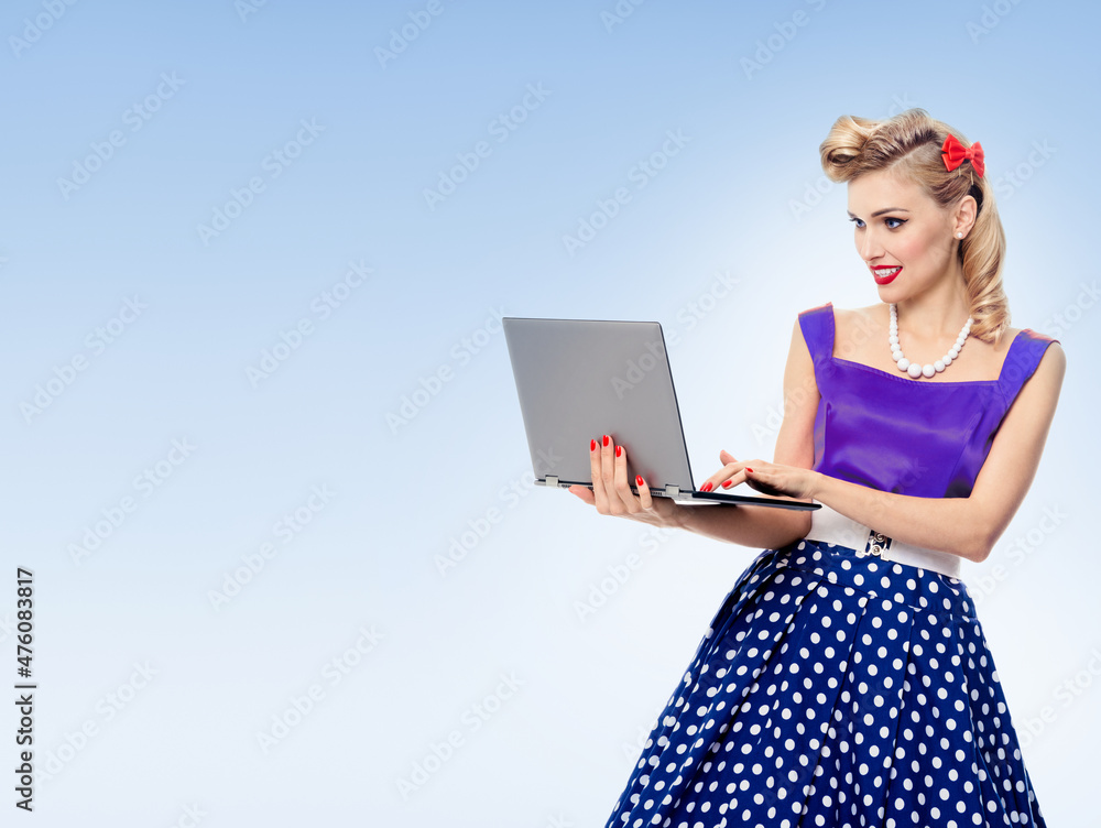 Portrait of beautiful woman holding laptop, dressed in pin-up style dress in polka dot, over light blue background. Blonde pinup girl posing in retro fashion and vintage concept studio shot. Portrait of beautiful woman holding laptop, dressed in pin-up style dress in polka dot, over light blue background. Blonde pinup girl posing in retro fashion and vintage concept studio shot.