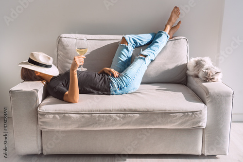 Young woman with glass of white wine resting on the sofa at home. Woman enjoying free time and lazy weekend. #476084651