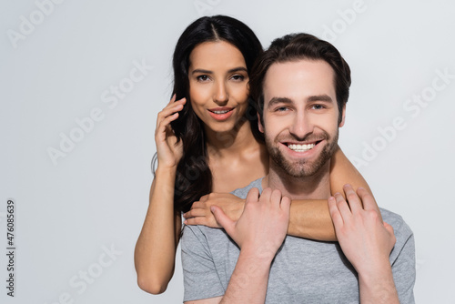 pretty brunette woman looking at camera while hugging bearded man in t-shirt isolated on grey.