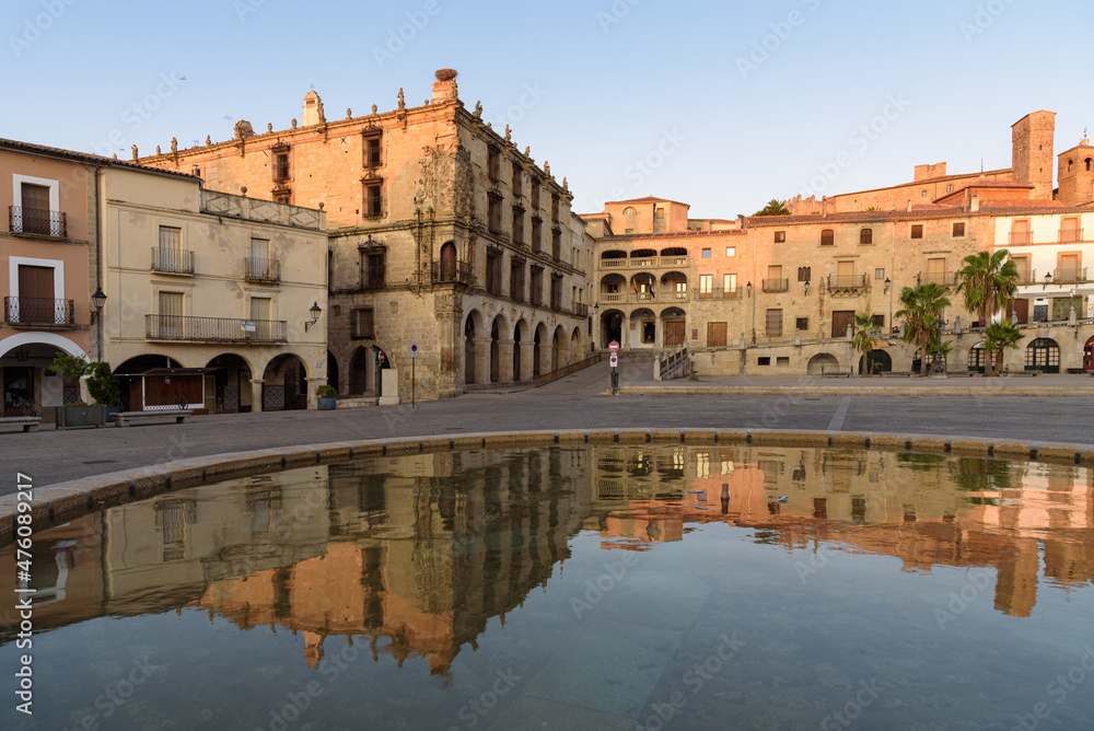 City landscape of the main square of the medieval city of Trujillo with its old stone buildings and arcades reflected in the water of the fountain at sunrise, Caceres, Extremadura, Spain
