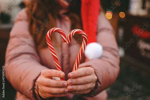 In the background, a young happy woman at the Christmas market smiling on a blurred background wearing a Santa Claus hat, shows a heart symbol made from red candy canes as a sign of support, love.