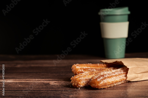 churros coming out of paper bag on wooden table. Mug of coffee on background