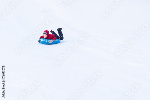 Smiling boy in winter clothes sliding on blue tubing in distance among white snow. Inflatable sleigh rides.
