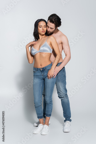 full length view of shirtless man and sexy woman in bra standing in jeans on grey.