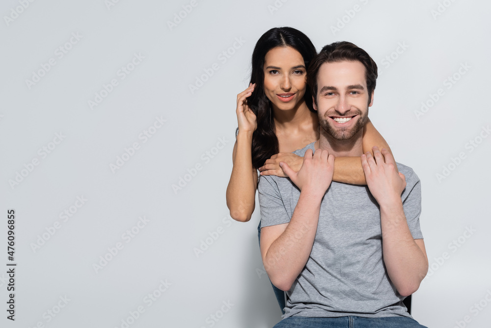 joyful man in t-shirt smiling at camera near seductive woman embracing him on grey. joyful man in t-shirt smiling at camera near seductive woman embracing him on grey.