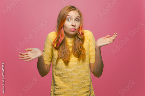 I don't know. Puzzled helpless young girl shrugging shoulders, spreading hands in uncertainty on pink studio background