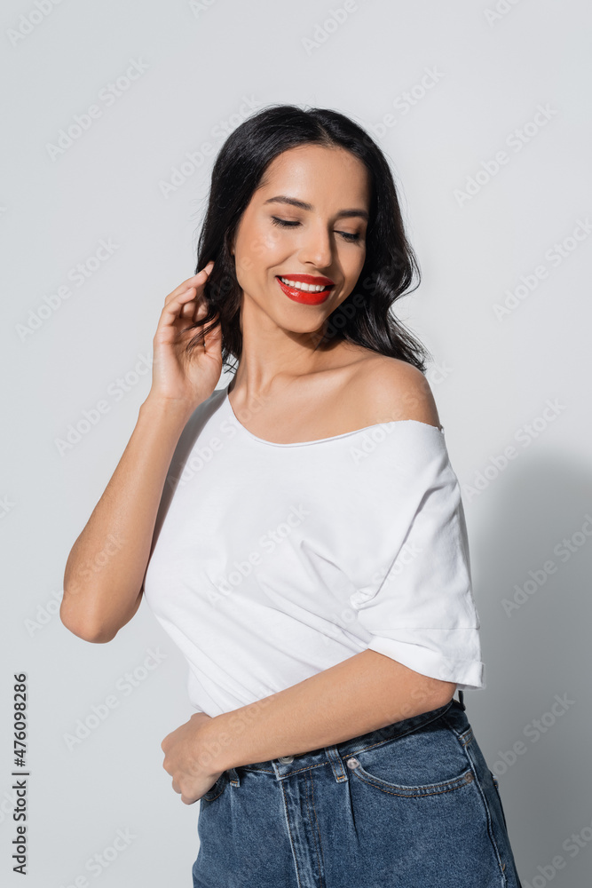 sensual and smiling woman in white t-shirt with naked shoulder touching hair while posing on grey. sensual and smiling woman in white t-shirt with naked shoulder touching hair while posing on grey.