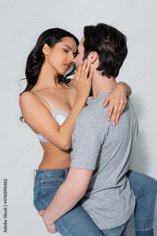 passionate woman in bra and jeans kissing man in t-shirt on grey.