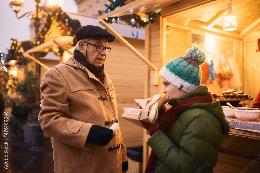 Senior man and his grandson eat in front of Christmas market stall during winter fair.