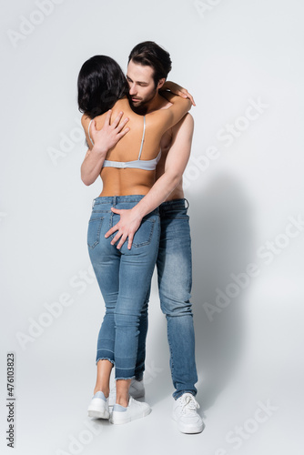 back view of sexy woman in jeans and bra near shirtless man hugging her on grey.