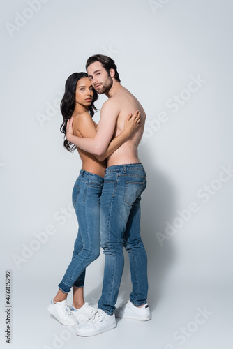 full length view of sexy couple in jeans and sneakers looking at camera while embracing on grey.