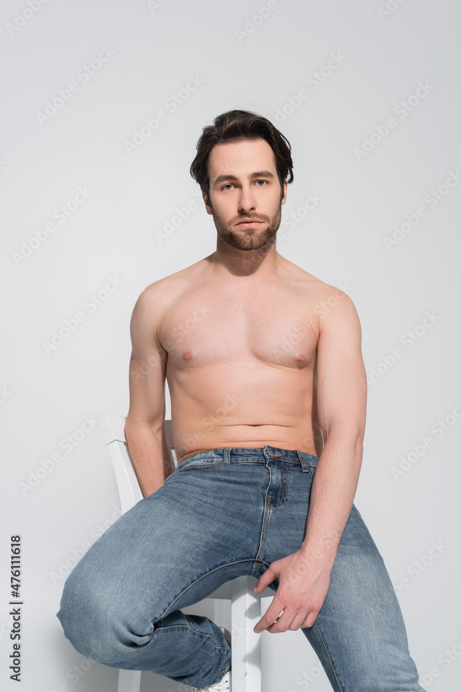 brunette shirtless man in jeans sitting on white chair and looking at camera on grey. brunette shirtless man in jeans sitting on white chair and looking at camera on grey.