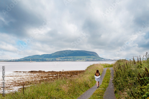 Teenager girl walking on a small country road with amazing view on the ocean and mountain and cloudy sky. County Sligo, Ireland. Knocknarea hill in the background. Travel and tourism concept photo