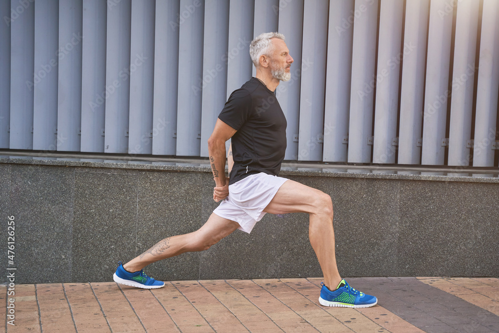 Confident sportive middle aged man in sports clothing stretching his legs, warming up and exercising outdoors Confident sportive middle aged man in sports clothing stretching his legs, warming up and exercising outdoors
