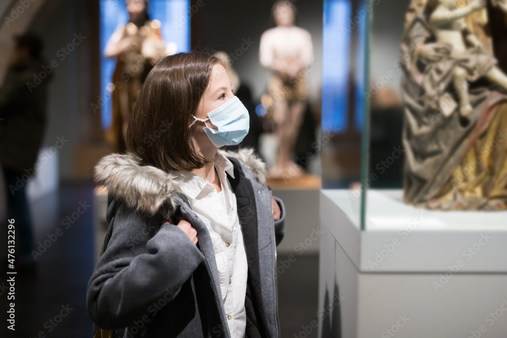 Positive tween schoolgirl in medical face mask observing with interest arts and crafts on exhibition in art gallery. New normal in coronavirus pandemic