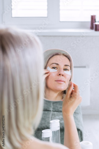 A beautiful young woman is taking care of her face, looking in the mirror applying eye patches. SPA. Facial care at home