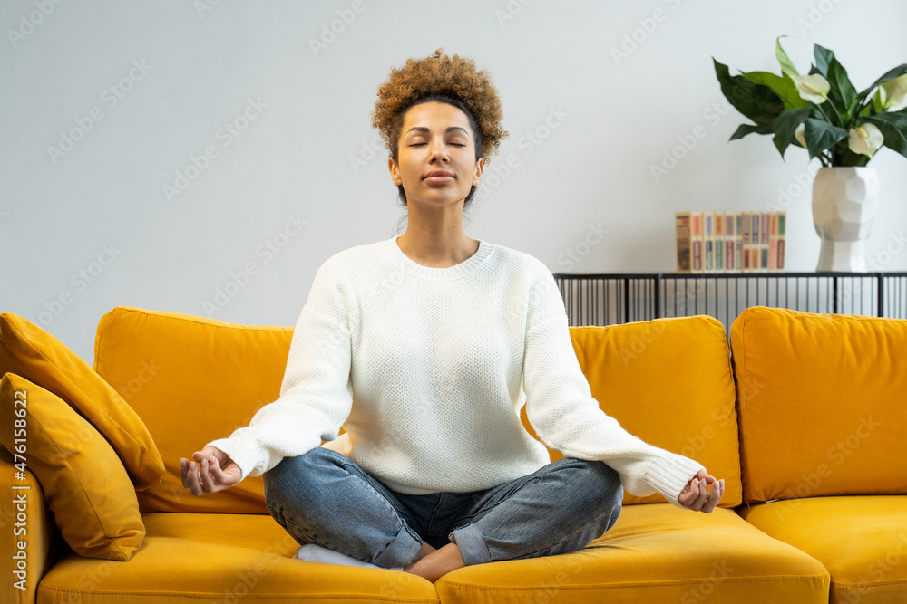 An African-American woman sitting on a comfortable sofa is doing yoga in the lotus position. Beautiful woman meditates on the couch at home An African-American woman sitting on a comfortable sofa is doing yoga in the lotus position. Beautiful woman meditates on the couch at home