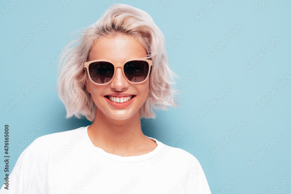 Portrait of young beautiful smiling female in trendy summer white clothes.Sexy carefree blond woman posing near blue wall in studio. Positive model having fun indoors. Cheerful and happy in sunglasses Portrait of young beautiful smiling female in trendy summer white clothes.Sexy carefree blond woman posing near blue wall in studio. Positive model having fun indoors. Cheerful and happy in sunglasses
