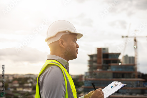 Inspector engineer man or architect with clipboard with white safety helmet in city construction site. Checking with checklist on rooftop building construction at capital.