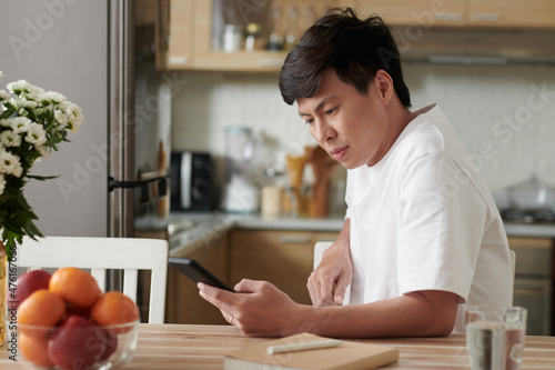 Frowning man sitting at kitchen table and reading notifications on smartphone