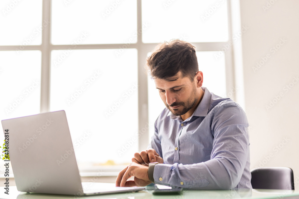 Portrait of businessman sitting at office while looking at his watch. Young professional getting ready for business meeting. Portrait of businessman sitting at office while looking at his watch. Young professional getting ready for business meeting.