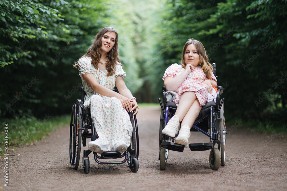 Two caucasian women in wheelchairs posing on camera among green city park. Female friends who live with disability having hairstyle and wearing beautiful summer dresses. Two caucasian women in wheelchairs posing on camera among green city park. Female friends who live with disability having hairstyle and wearing beautiful summer dresses.
