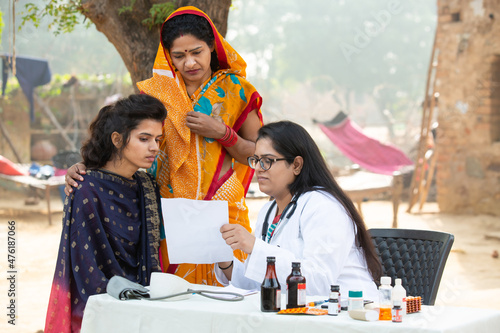 Young Indian adult girl with her mother getting proscription medicine by female doctor at village outdoor at government camp, woman examine by medical person, Rural India healthcare.