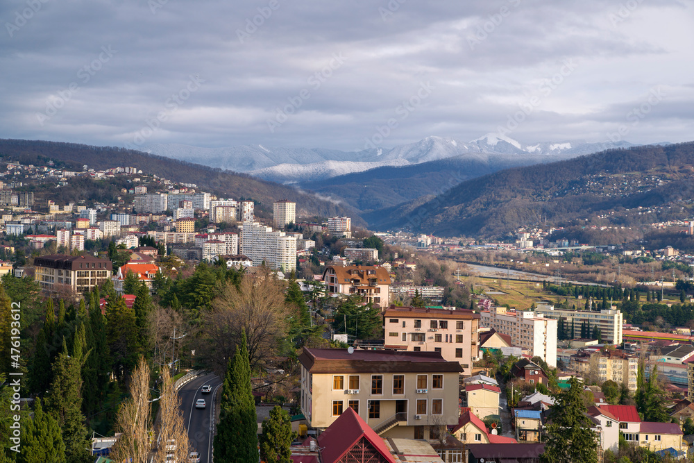 View of the city from a height against the background of snow-covered mountains and cloudy sky. Beautiful panorama of Sochi in winter.