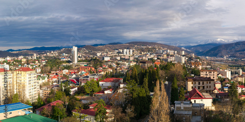 View of the city from a height against the background of snow-covered mountains and cloudy sky. Beautiful panorama of Sochi in winter.