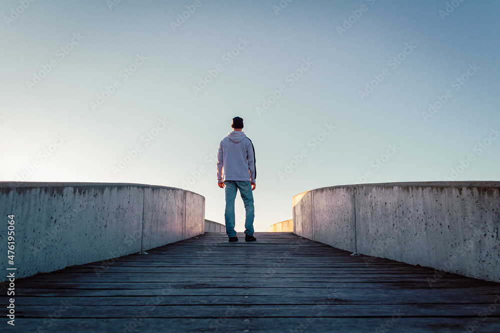 Young caucasian man in jeans, hat and hoodie standing on concrete bridge. Rear view male pose in city environment and clear sky