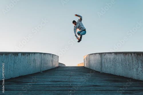 Young caucasian man in jeans and hoodie jumping on concrete bridge. Mid air parkour pose in city environment and clear sky photo