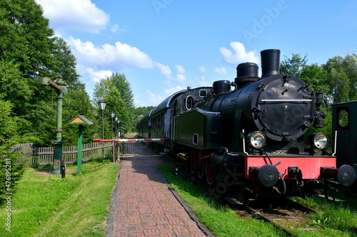 A close up on an old yet still used steam locomotive with some compartments standing on a platform next to a pavement and some trees being a part of a lush forest or moor seen in Poland in summer
