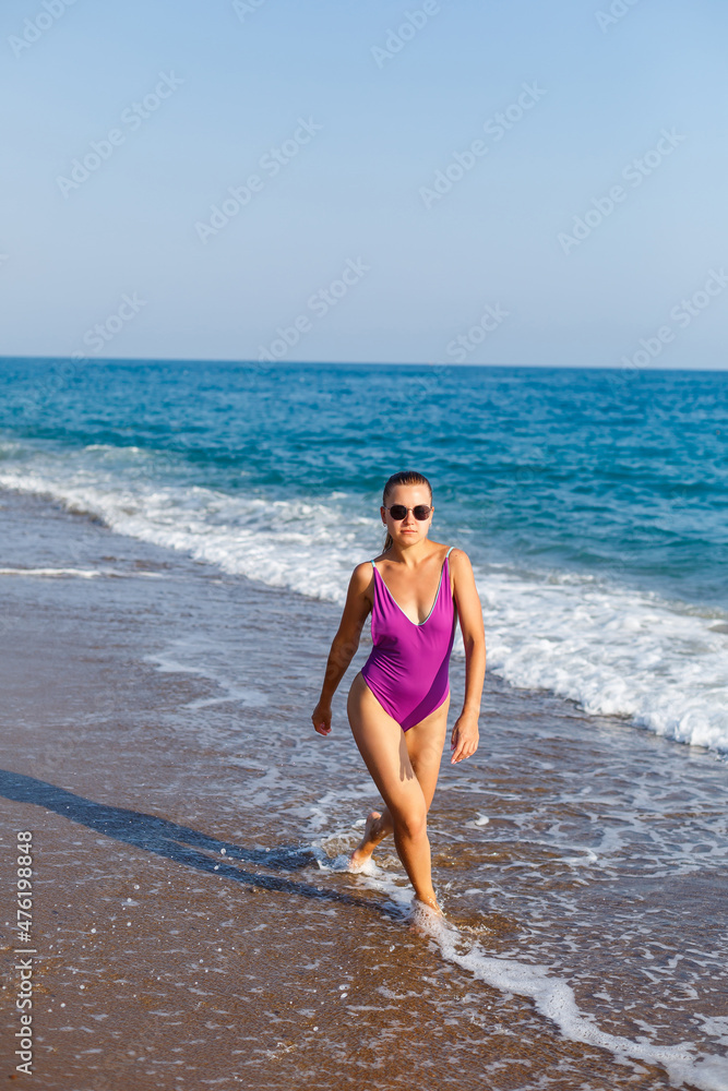 A young beautiful woman with a toned body in a bright swimsuit walks along the sandy beach. Summer vacation at the sea. Mediterranean Turkish Sea. Selective focus