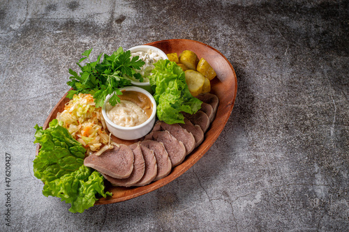 Serving a dish from the restaurant menu. Sliced beef tongue with pickles, cabbage, sauces and herbs on a plate against a gray stone table, a delicious appetizer