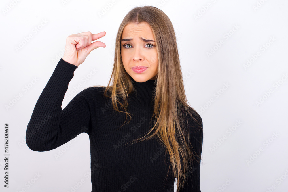 Upset Young caucasian girl wearing black turtleneck over white background shapes little gesture with hand demonstrates something very tiny small size. Not very much Upset Young caucasian girl wearing black turtleneck over white background shapes little gesture with hand demonstrates something very tiny small size. Not very much