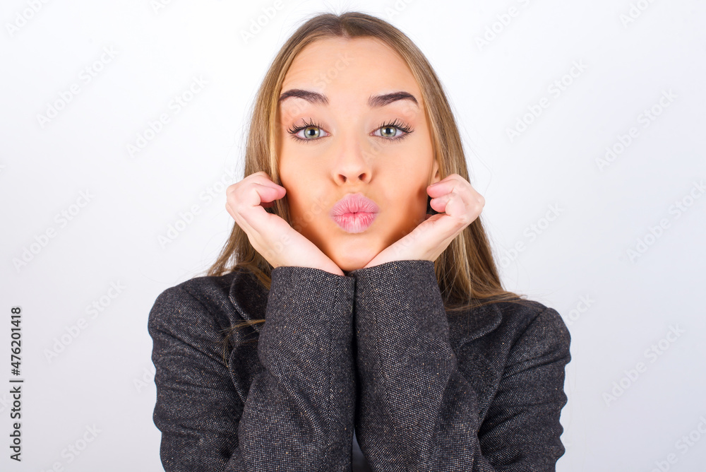 Young business woman wearing jacket over white background with surprised expression keeps hands under chin keeps lips folded makes funny grimace Young business woman wearing jacket over white background with surprised expression keeps hands under chin keeps lips folded makes funny grimace
