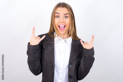 Young business woman wearing jacket over white background makes rock n roll sign looks self confident and cheerful enjoys cool music at party. Body language concept.