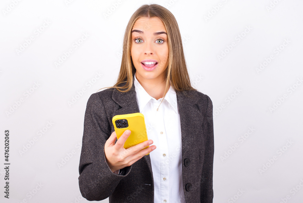 Young business woman wearing jacket over white background holds mobile phone in hands and rejoices positive news, uses modern cellular Young business woman wearing jacket over white background holds mobile phone in hands and rejoices positive news, uses modern cellular