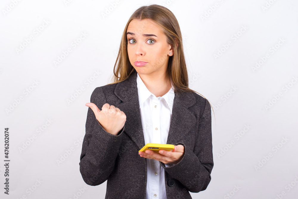 Young business woman wearing jacket over white background points thumb away and shows blank space aside, holds mobile phone for sending text messages. Young business woman wearing jacket over white background points thumb away and shows blank space aside, holds mobile phone for sending text messages.
