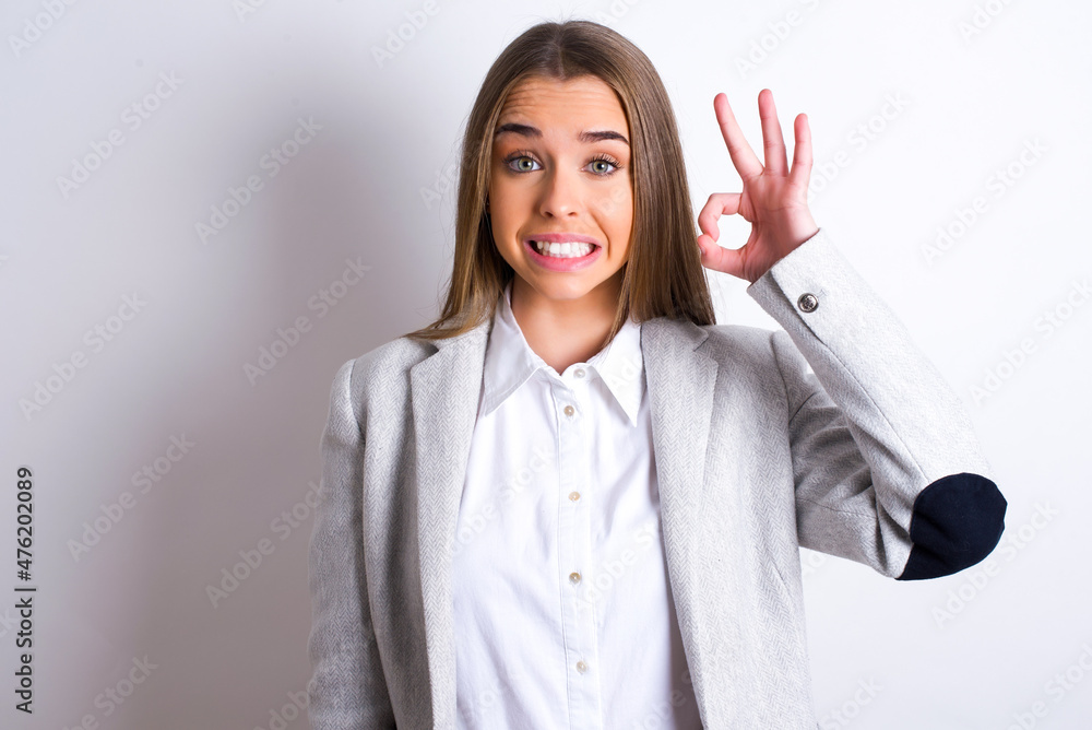 Young business woman wearing jacket over white background hold hand arm okey symbol toothy approve advising novelty news Young business woman wearing jacket over white background hold hand arm okey symbol toothy approve advising novelty news