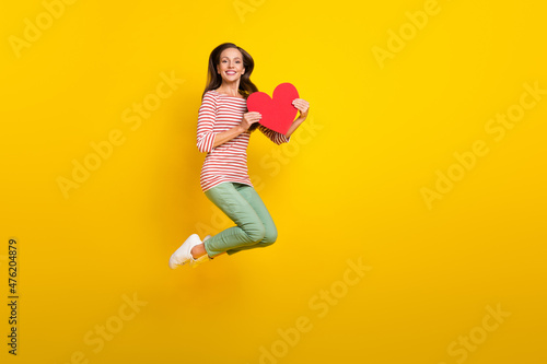 Photo of charming pretty young woman wear striped shirt jumping high holding large heart empty space isolated yellow color background