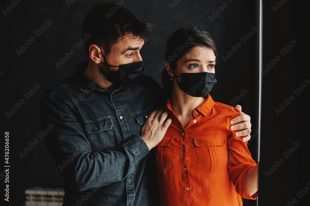 Scared couple with face masks standing next to window and looking outside. Lockdown during corona virus concept. Scared couple with face masks standing next to window and looking outside. Lockdown during corona virus concept.
