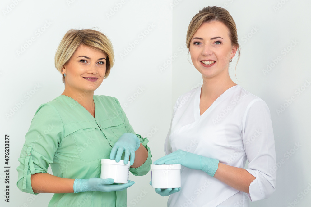 Obraz premium Two cosmeticians with jars of wax for depilation smiling against a white background. Natural product for hair removal. Copy space