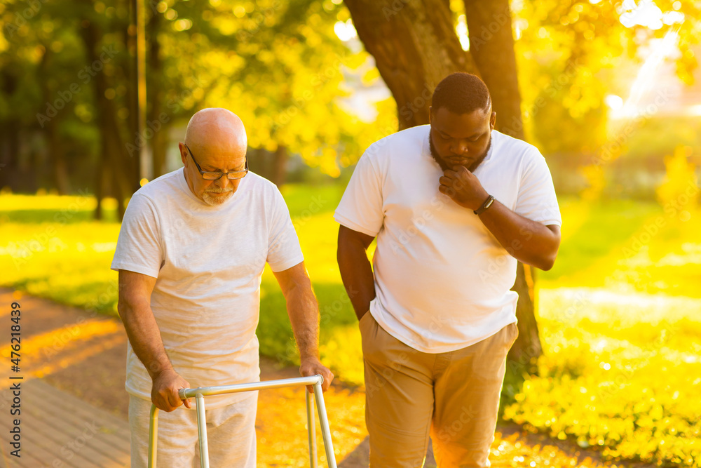 Obraz premium Caregiver is teaching old man to walk with walker. Professional nurse and patient walking outdoor at sunset. Assistance, rehabilitation and health care.